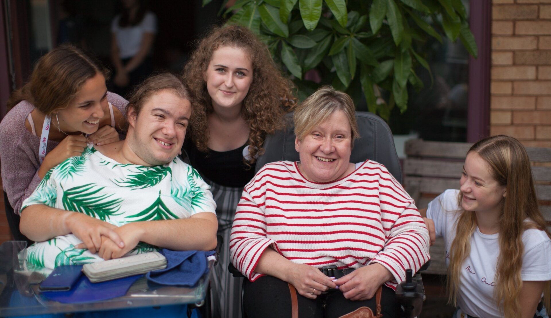 Two Revitalise guests in wheelchairs supported by three friendly and smiling young female volunteers