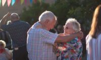Elderly woman smiling at the man she is dancing with on a sunny day during a garden party at Revitalise Jubilee Lodge