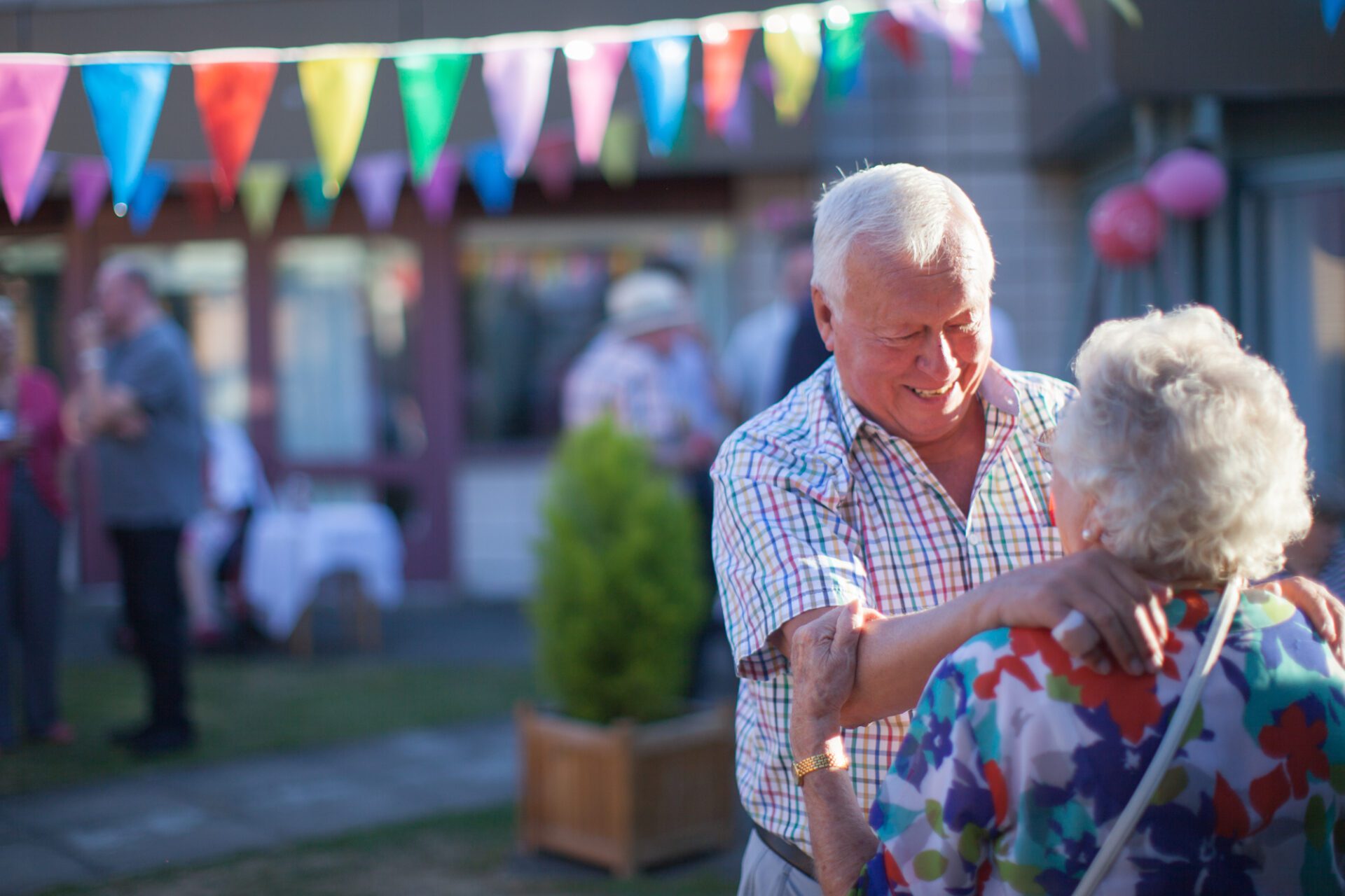 An elderly man smiling at the woman he's dancing with on a sunny dayin the courtyard of Revitalise Jubilee Lodge