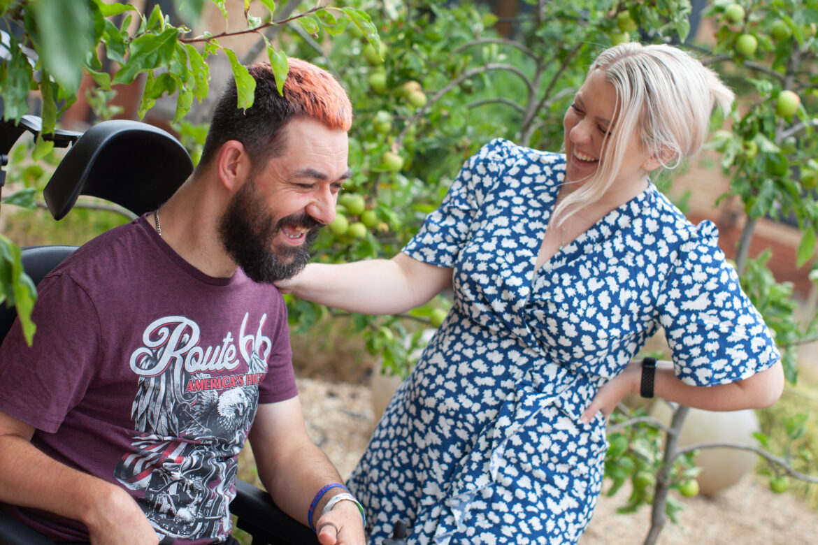 Young man with pink hair in a wheelchair laughing with a young woman with blonde hair in a blue dress in the courtyard at Revitalise Jubilee Lodge