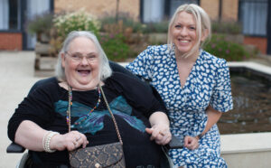 Older female Revitalise guest sitting with young woman in blue dress, both smiling, in the courtyard at Jubilee Lodge