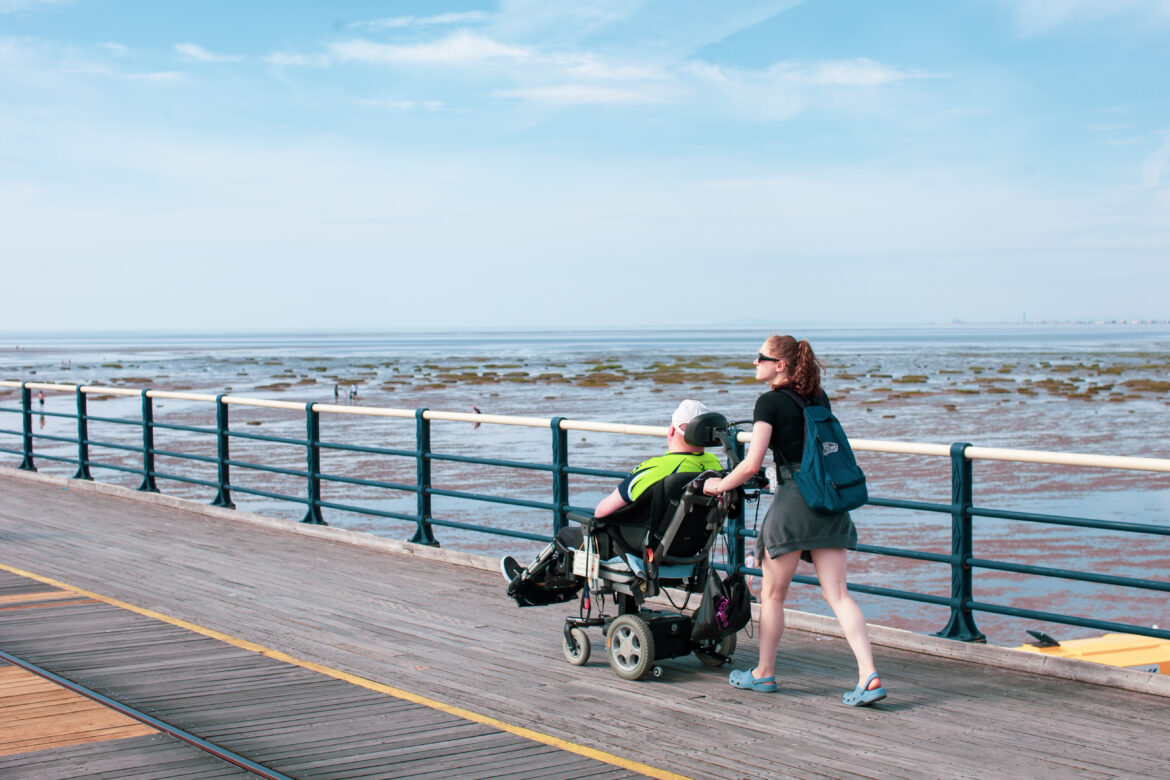 Young woman pushing a man in a wheelchair along the pier in Southport during a Revitalise respite holiday