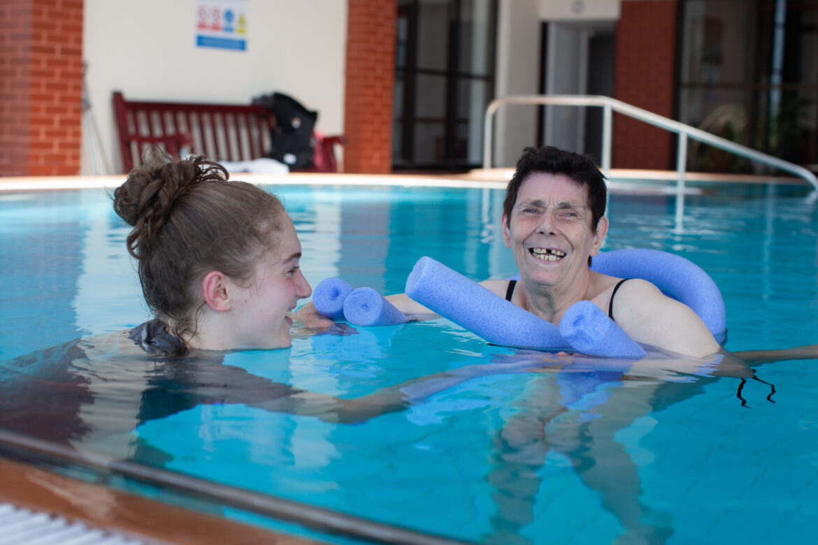 Woman floating with pool noodle in the Revitalise Sandpipers hydrotherapy pool with a volunteer