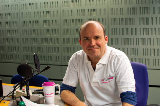Actor Rory Kinnear smiling in the sound booth at the BBC recording studio, wearing a white Revitalise polo shirt.