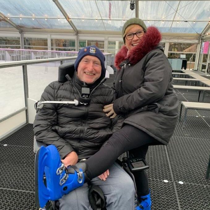 Man in a wheelchair with a woman. Both are bundled up and enjoying an ice skating excursion and smiling at the camera.