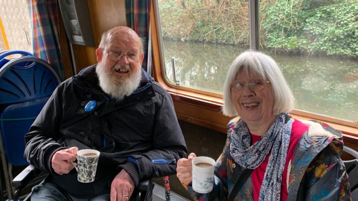 Image of two Revitalise guests sitting inside a boat, while enjoying a canal tour in London. Both guests are holding a warm drink and are smiling at the camera.