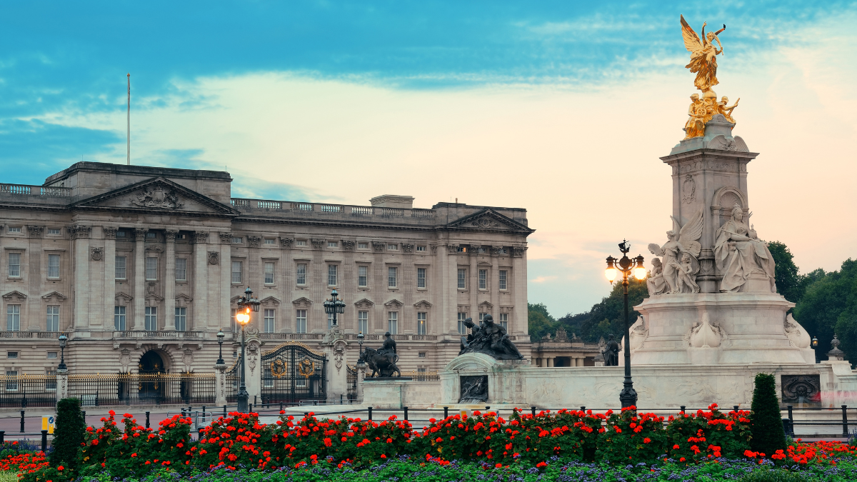 A long shot of Buckingham Palace in London. The Queen Victoria memorial and a garden filled of red flowers can also be seen in the foreground.