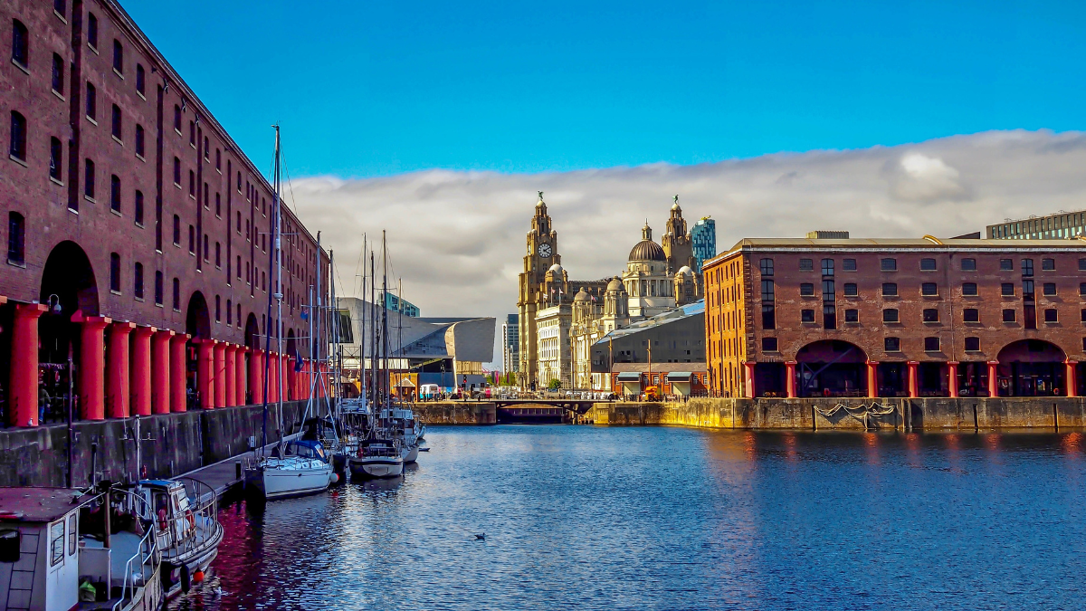 A view of the Liverpool waterfront as seen from The Royal Albert Dock. Boats can be seen docked on the left side of the water body. In the background, Liverpool's historic and cultural buildings such as Tate and Maritime Museum are visible.