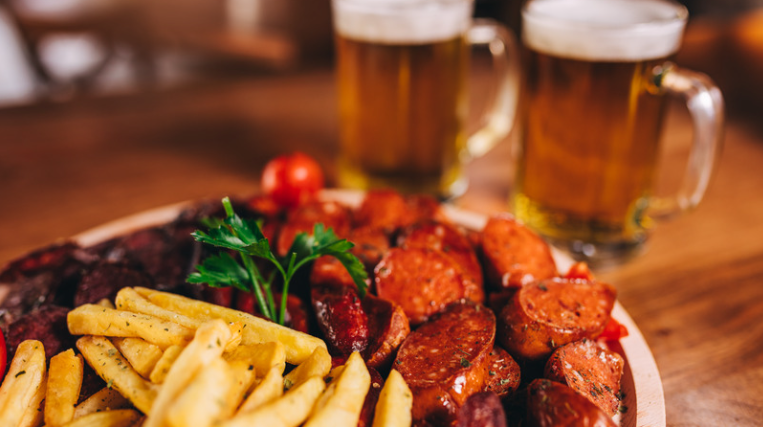 Image of a plate stacked with sliced sausages, fries and salad. Two mugs filled with beer are also visible in the background.