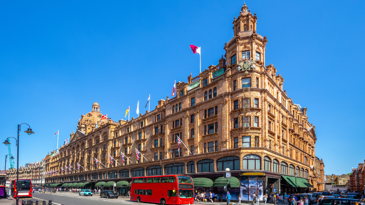 An image of the Harrods in London taken from street. The Edwardian baroque design and curved entrance windows that this building is famous for, are visible in the image. The image also shows a red London bus passing through in front of the building.