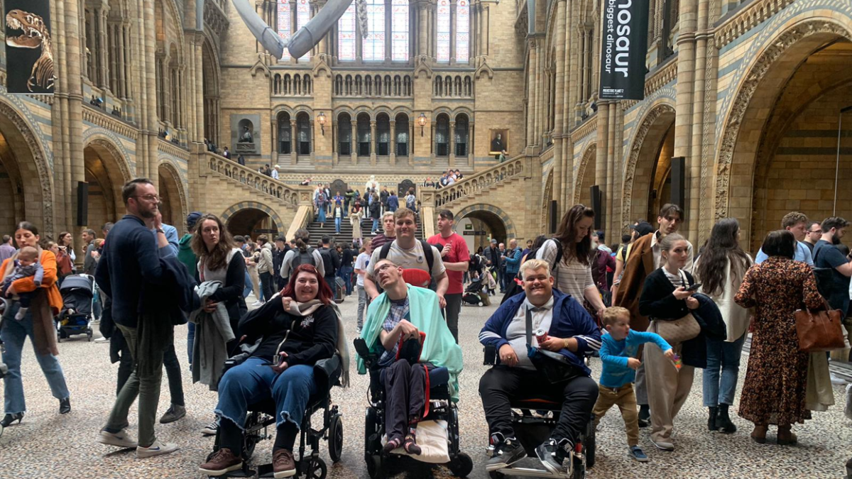 Guests and volunteers from Revitalise Jubilee Lodge can be seen out on an excursion to the Natural History Museum in London. They are in a big indoor crowded courtyard large windows. Skeleton of a giant whale is hanging from the ceiling.