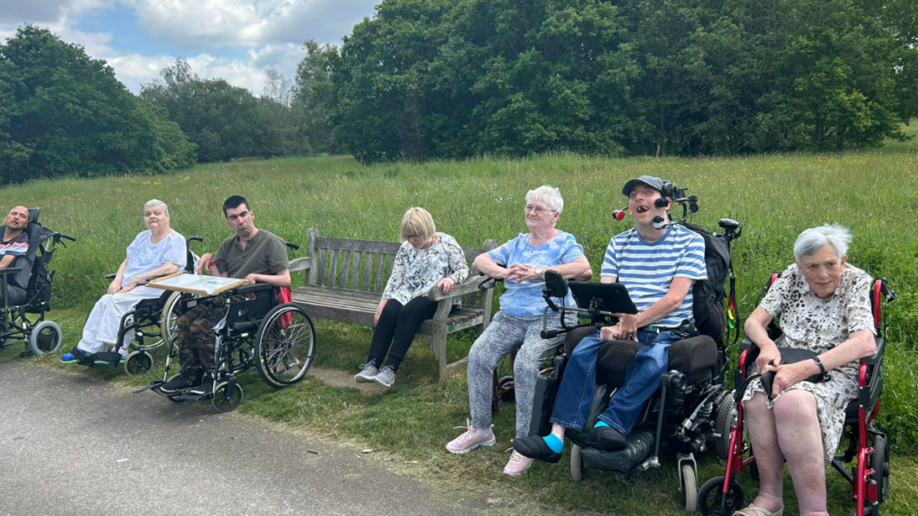 Guests at Jubilee Lodge holiday centre out on an excursion to a park. The guests are using wheelchair and have lined up for the picture. Lush green trees and blue sky with puffy clouds can be seen in the background.