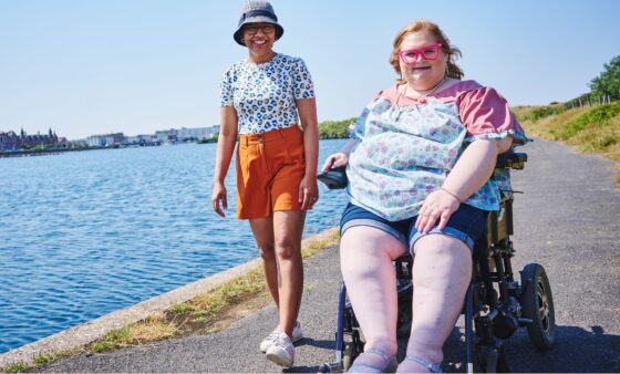 Revitalise Sandpipers guest in electric wheelchair on a walk around Southport's Marine Lake on a sunny day with a volunteer