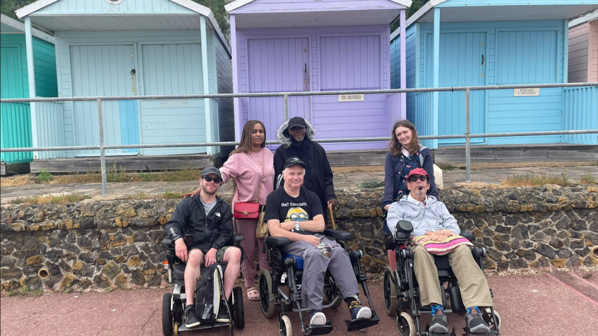 Image of three guests at Revitalise out on an excursion to seaside along with three volunteers. The guests are using wheelchair and smiling at the camera. Blue and purple pastel beach huts can be seen in the background.