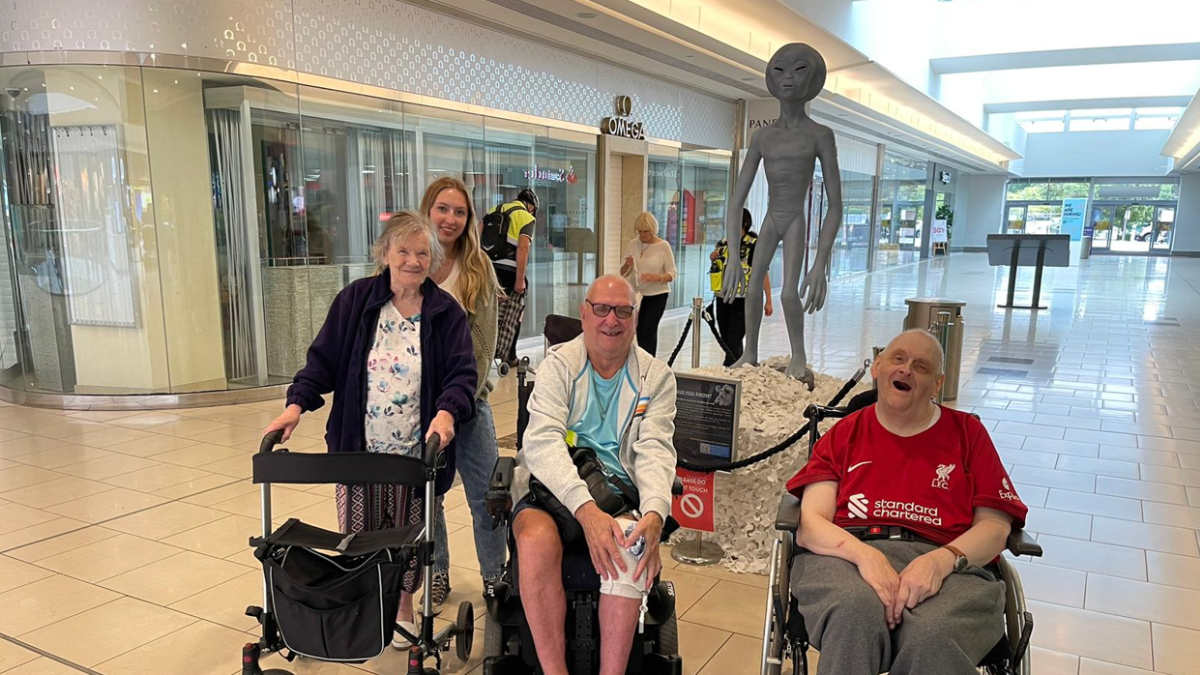Image of three Revitalise guests and a volunteer at a trip to a shopping mall in London. Two of the guests are using a wheelchair and one of them is using a mobility aid. A sculpture of an alien like figure can be seen in the background.