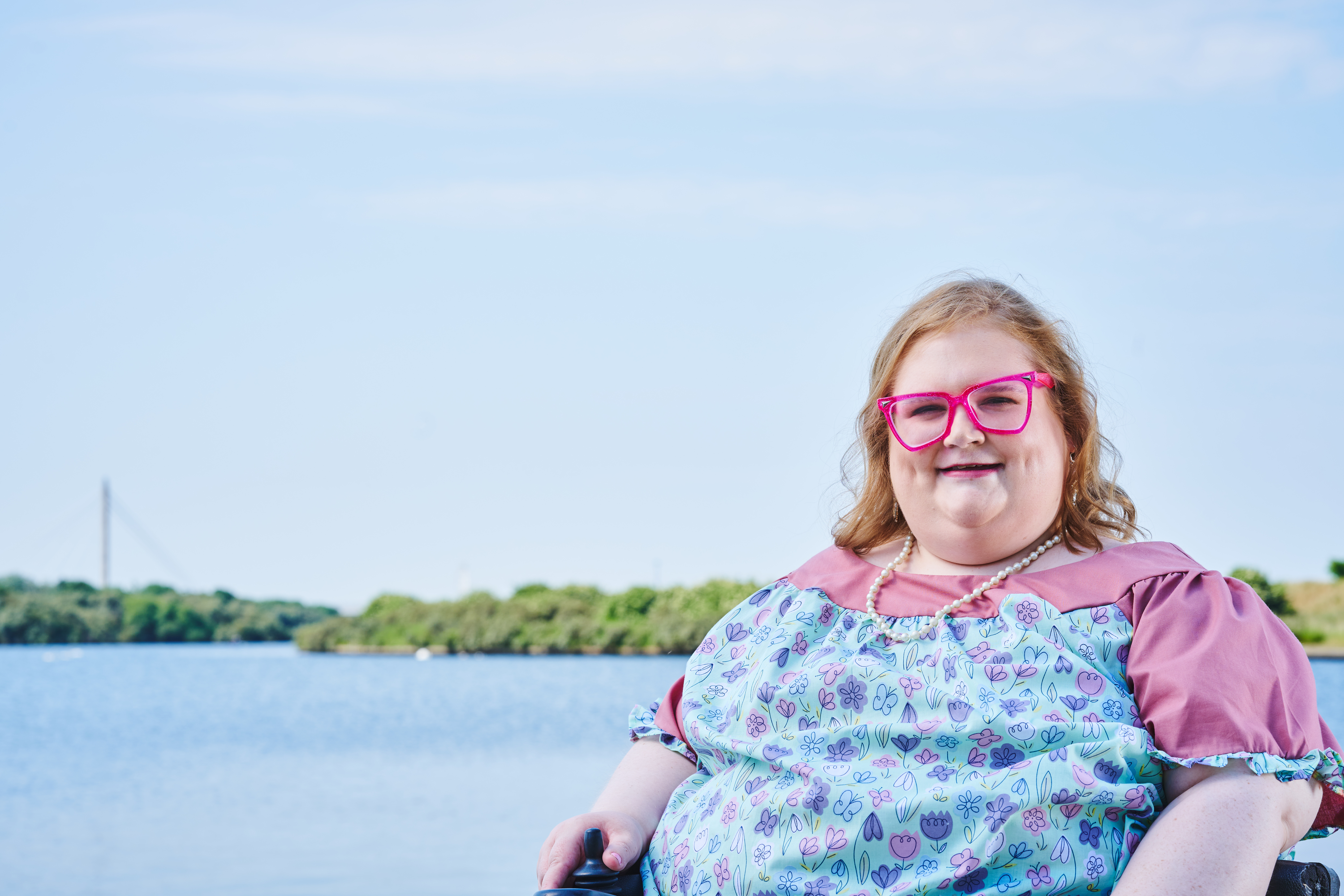 Woman in an electric wheelchair and Revitalise Sandpipers guest with bright pink glasses enjoying a sunny day in front of Southport's Marine Lake