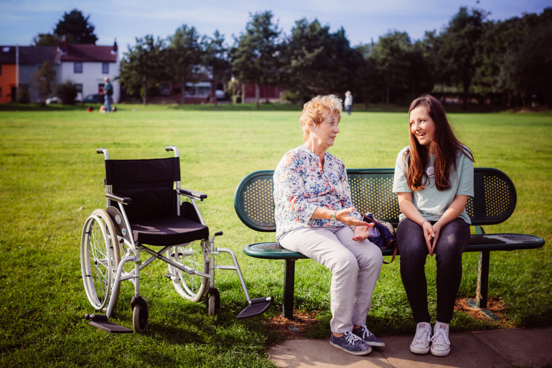 Two women sitting on a park bench on a sunny day. The older of the women has a wheelchair next to the bench.