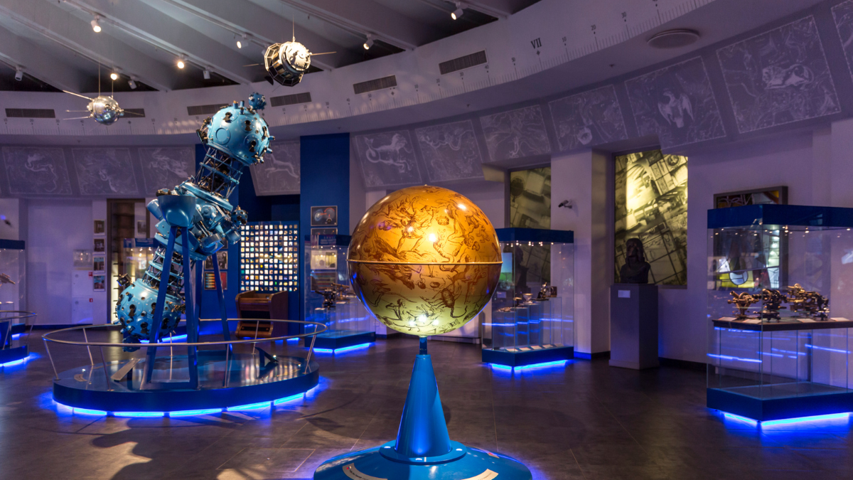 Image of the astronomy exhibit of the Science Museum in London. The image focuses on a large yellow-coloured model of earth balancing on a blue stand. Replica of large and small satellites can be seen in the background.