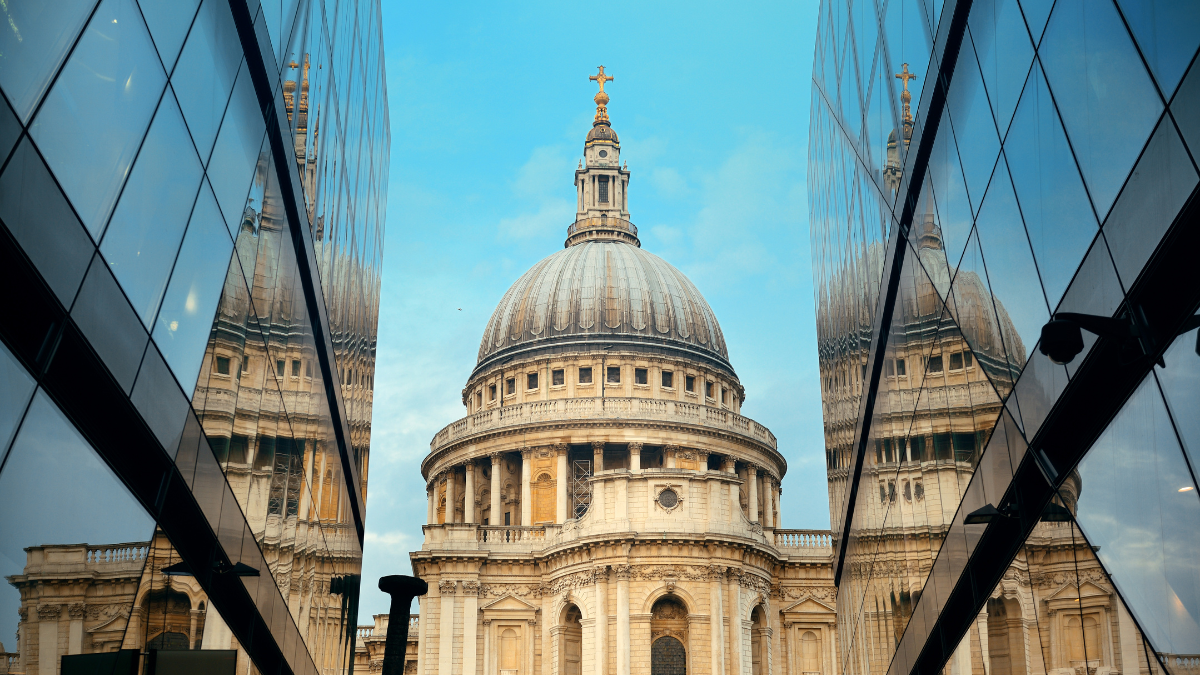 Image shows a long shot view of St Paul's Cathedral taken from the One New Change building on Bread Street. The street has glass buildings on both sides which are reflecting St Paul's Cathedral and the bright blue sky