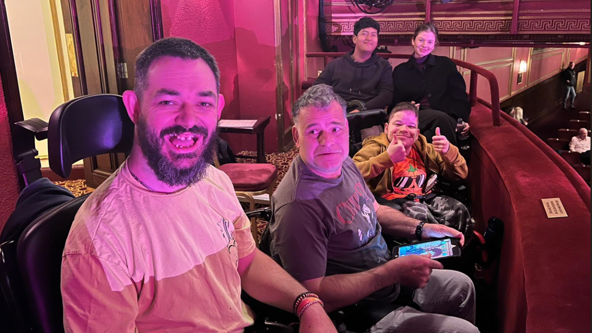 Three Revitalise guests enjoying a live theatre show at London's West End. Image has a pink hue, possibly from the lights of the theatre. The guests are smiling at the camera, with one of them giving thumbs up to the performance.