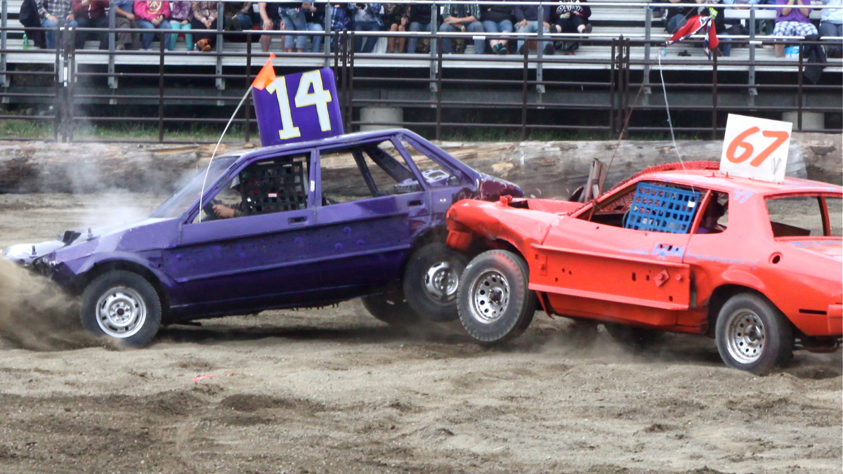 Image of two cars participating in banger racing. A red car with the number 67 on top is crashing into a purple car with the number 14 on top, in a muddy stadium.