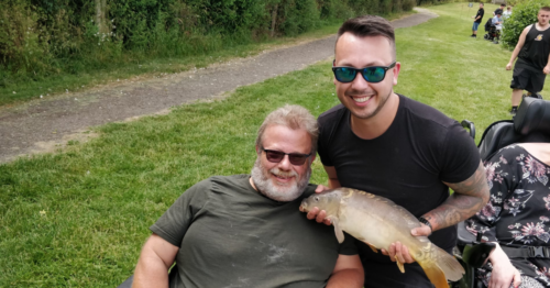 Image of a guest from Revitalise Jubilee Lodge with a volunteer from The Prince's Trust enjoying a fishing excursion. The guest is using a wheelchair and the volunteer is holding a fish. Both of them are wearing sunglasses and smiling at the camera.