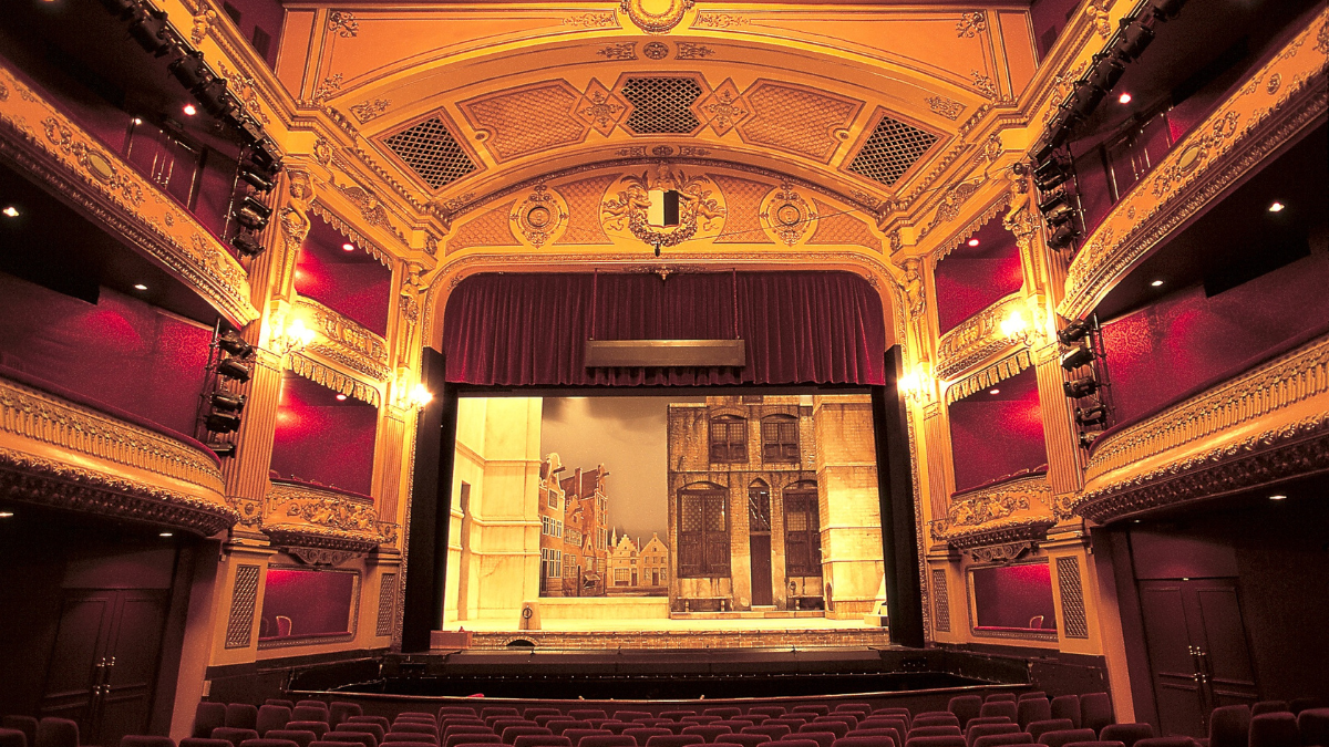 A wide angle image of an old empty theatre with red stage curtains and dramatic lights.